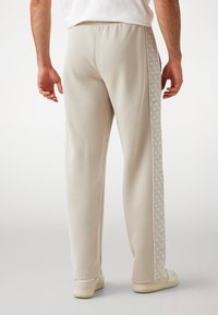 Helle beige Jogginghose aus weichem Material, mit glatter Textur, elastischem Bund und einem gemusterten Seitenstreifen.