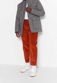 Giacca piumino grigia con chiusura a zip e bottoni, abbinata a pantaloni in velluto a coste arancioni e sneakers bianche. La giacca presenta tasche frontali.