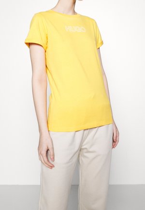 Osoba nosící žluté tričko s krátkým rukávem s nápisem "HUGO" a béžové kalhoty, stojící před jednobarevným bílým pozadím.