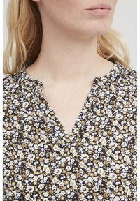Bloemenprint top met een marineblauwe achtergrond, voorzien van kleine witte en gele bloemen, een geknipte halslijn en zachte plooien op de schouders.