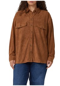 Person in einem lockeren braunen Wildlederhemd mit Knöpfen und zwei Brusttaschen, kombiniert mit blauen Jeans, vor einem weißen Hintergrund.