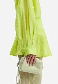 Borsa verde chiaro di forma rettangolare, con chiusura a zip e tracolla removibile. Indossata sotto una blusa verde brillante e con texture.