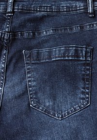 Detailní záběr na zadní kapsu tmavě modrých džín, která vyniká švy a hladkým, texturovaným povrchem látky.