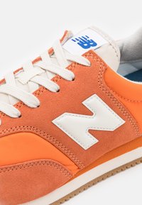 La basket orange présente une tige en daim et synthétique, des accents blancs, des lacets ronds et une semelle en gomme texturée. Le logo est bien visible.