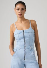 Νεαρή γυναίκα με σκούρα μαλλιά και σκουλαρίκια κρίκους, φοράει ανοιχτό μπλε ολόσωμη φόρμα τζιν με φερμουάρ μπροστά, τα χέρια στις τσέπες, στεκόμενη μπροστά σε απλό φόντο.