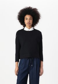 Black knit sweater with three-quarter sleeves, featuring button accents on shoulders. Paired with loose-fitting blue denim pants.