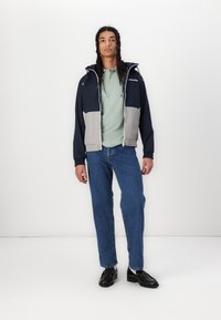Eine navyblaue und graue Windjacke mit Kapuze, Reißverschluss vorne und elastischen Bündchen, getragen über einem hellgrünen Poloshirt und kombiniert mit blauen Jeans und schwarzen Schuhen.