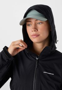 Femme ajustant la capuche d'une veste noire zippée Swedemount, portant une casquette de baseball bleu clair en dessous.