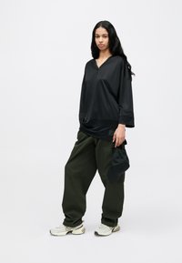 Μαύρο μακρυμάνικο τοπ με V-λαιμόκοψη και οριζόντια υφή, συνδυασμένο με φαρδιά ελιά παντελόνια και ανοιχτόχρωμ α αθλητικά παπούτσια, κρατώντας μαύρο καπέλο.