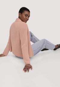Strickjacke in zartem Blush-Pink mit strukturiertem Muster, offener Vorderseite und langen Ärmeln. Kombiniert mit hellblauen Jeans und dunklen Schuhen.