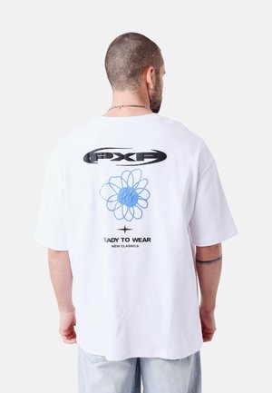 Hombre con cabello corto y tatuaje, vistiendo una camiseta blanca de gran tamaño con un diseño gráfico negro y azul y texto en la parte trasera.