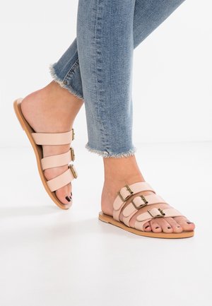 Feet wearing light pink triple-strap slide sandals with gold buckles, paired with light blue frayed hem skinny jeans on a white floor.