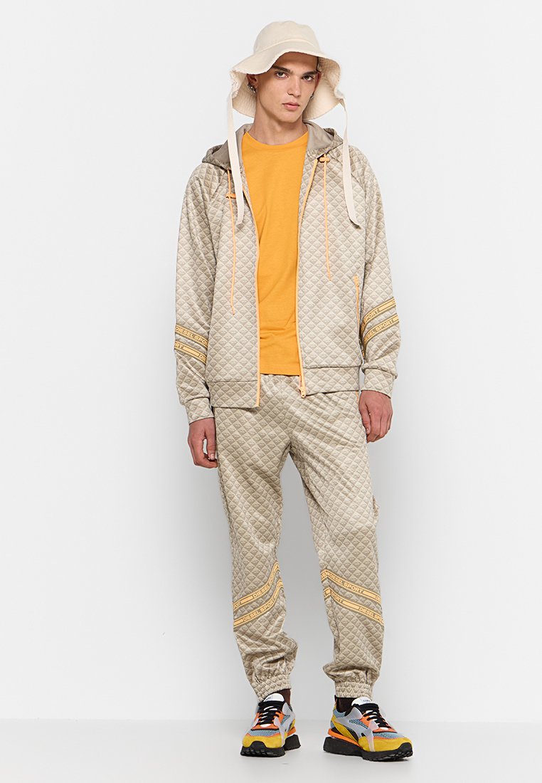 Set giacca e pantaloni trapuntati beige con dettagli arancioni, abbinato a una maglietta gialla e un cappello a secchiello beige. Sneakers multicolori.