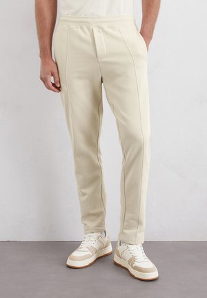 Cremfarbene Fleece-Jogginghosen mit schmalem Schnitt, elastischem Bund und Kontrastnähten, kombiniert mit weißen und beigen Turnschuhen.