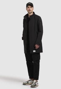 Abrigo negro hasta la rodilla con un cuello alto, tejido texturizado y bolsillos laterales. Combinado con pantalones cargo negros y zapatillas en blanco y negro.