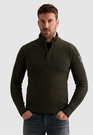Homme portant un pull texturé vert foncé à demi-fermeture éclair avec manches côtelées et jean, debout les mains dans les poches devant un fond uni.