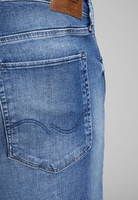 Denim jeans feature a blue wash with whiskering, a back pocket with curved stitching, and a brown leather patch at the waistband.