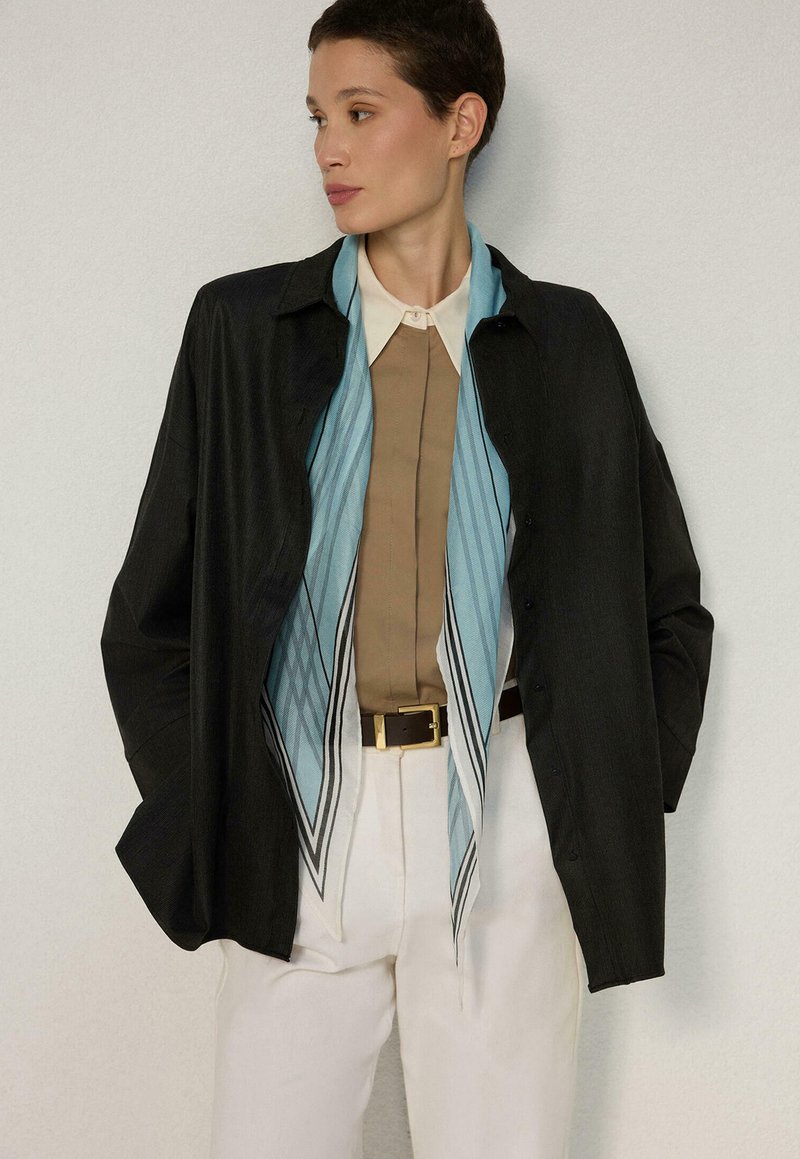 Femme aux cheveux courts portant une veste noire boutonnée, une écharpe rayée bleu clair, une chemise beige, un pantalon blanc et les mains dans les poches, regardant sur le côté.