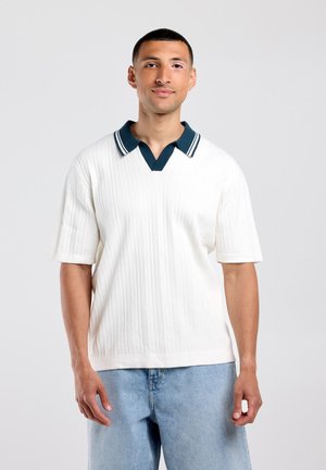 Man met kort donker haar die een witte gestructureerde korte mouwtrui met een donkere kraag en lichtblauwe spijkerbroek draagt, staand tegen een effen achtergrond.