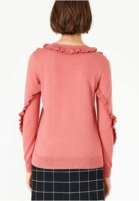 Růžový svetr s rýhovanými akcenty na ramenou a rukávech. Měkká textura, přiléhavý design a žebrovaný lem. Nesený přes kostkovanou sukni.