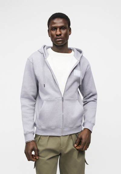 Άνδρας με σκουρόχρωμο δέρμα που φοράει ανοιχτό γκρι φούτερ με φερμουάρ πάνω από λευκό πουκάμισο και ελιά πράσινο παντελόνι, στέκεται μπροστά σε απλό λευκό φόντο.