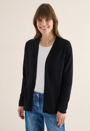 Femme souriante aux cheveux bruns porte un cardigan noir ouvert sur un haut blanc et un jean bleu, debout devant un fond clair uni.
