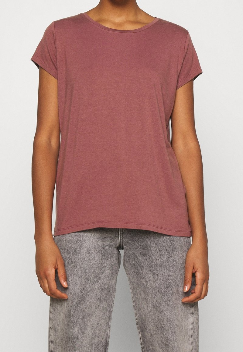 Personne portant un t-shirt mauve uni à manches courtes et un jean gris clair délavé, debout devant un fond uni.