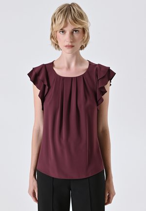 Donna bionda che indossa una blusa senza maniche di color bordeaux con spalle arricciate e pantaloni neri, su uno sfondo chiaro e uniforme.