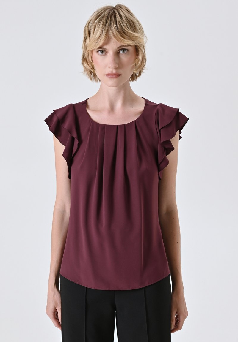 Blonde Frau, die eine bordeauxfarbene ärmellose Bluse mit Rüschenschultern und schwarze Hosen vor einem schlichten hellen Hintergrund trägt.