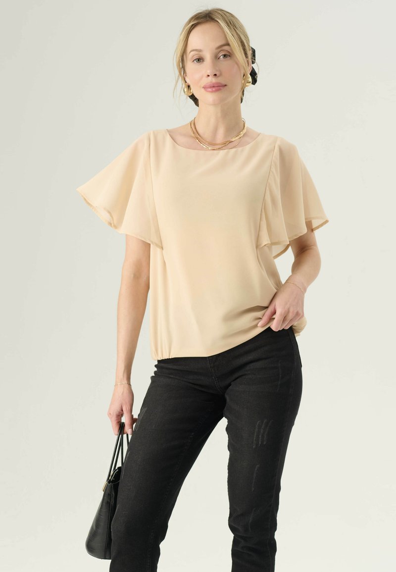 Beige chiffon top met korte vlinder mouwen en een ronde hals, gecombineerd met zwarte skinny jeans. Zwarte handtas in de hand, minimalistisch ontwerp.