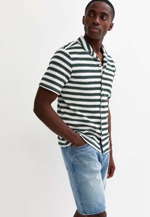 Homme portant une chemise à manches courtes rayée verte et blanche et un short en denim bleu clair, debout avec une main dans la poche.