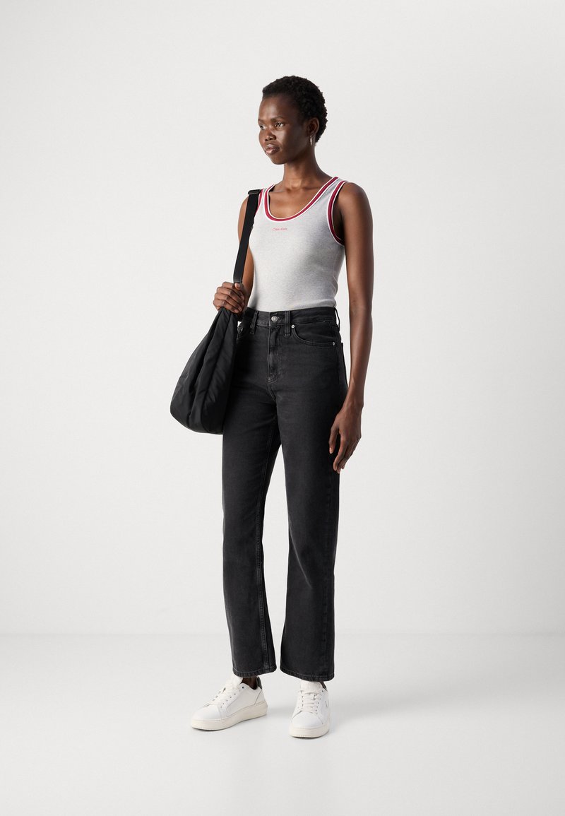 Frau in einem weißen Trägershirt mit rotem Saum, schwarzen Jeans und weißen Sneakers, die eine schwarze Schultertasche trägt und vor einem schlichten Hintergrund steht.