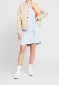 Chaqueta bomber acolchada beige con botones a presión, combinada con un vestido envolvente azul claro con cinturilla ajustada y zapatillas blancas con calcetines tobilleros.