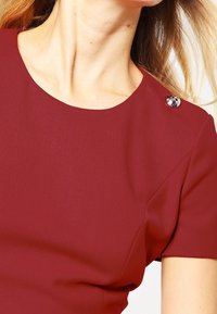 Il vestito bordeaux presenta un col rotondo con un accento di bottone argento sulla spalla e maniche corte, realizzato in un tessuto liscio.
