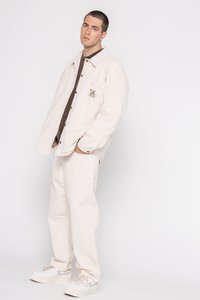 Witte oversized shirtjas met knopen en een voorzak, gelaagd over een bruine shirt. Crème-kleurige wijde broek en chunky beige sneakers.
