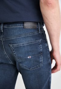 Mörkblå denimjeans med texturerad yta. Funktioner inkluderar en bakläderlapp märkt "JEANS" och en liten logotyp på fickan.