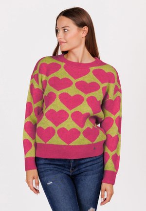 Frau in grünem Pullover mit rosa Herzmustern und blauen zerrissenen Jeans, die nach links schaut, vor einem schlichten Hintergrund.