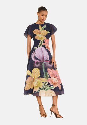 Žena nosi námornícky modré midi šaty s veľkými žltými, ružovými a fialovými kvetinovými potlačami a s voľnými rukávmi, skombinované s hnedými sandálmi na podpätku s remienkami.
