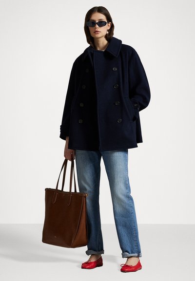 Marineblauw dubbelbreasted jas met grote knopen, blauwe jeans, rode platte schoenen en een bruin leren schoudertas. Minimalistische zonnebril maakt de look compleet.