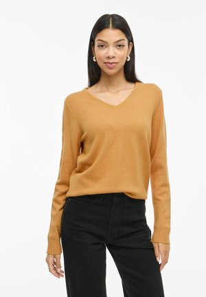 Femme portant un pull col en V couleur camel et un pantalon noir, debout dans une posture détendue devant un fond blanc uni.