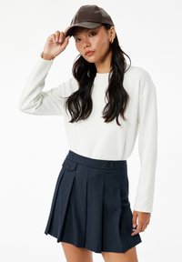 Weißer Pullover mit langen Ärmeln, marineblumiger Faltenrock und einer braunen Lederkappe. Der Rock hat Knopfdetails und eine vertikale Struktur.
