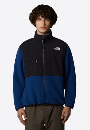 Giacca in pile nera e blu con colletto alto; tasca sul petto con zip; logo sul lato sinistro del petto; tessuto texturizzato; vita e polsini elasticizzati.
