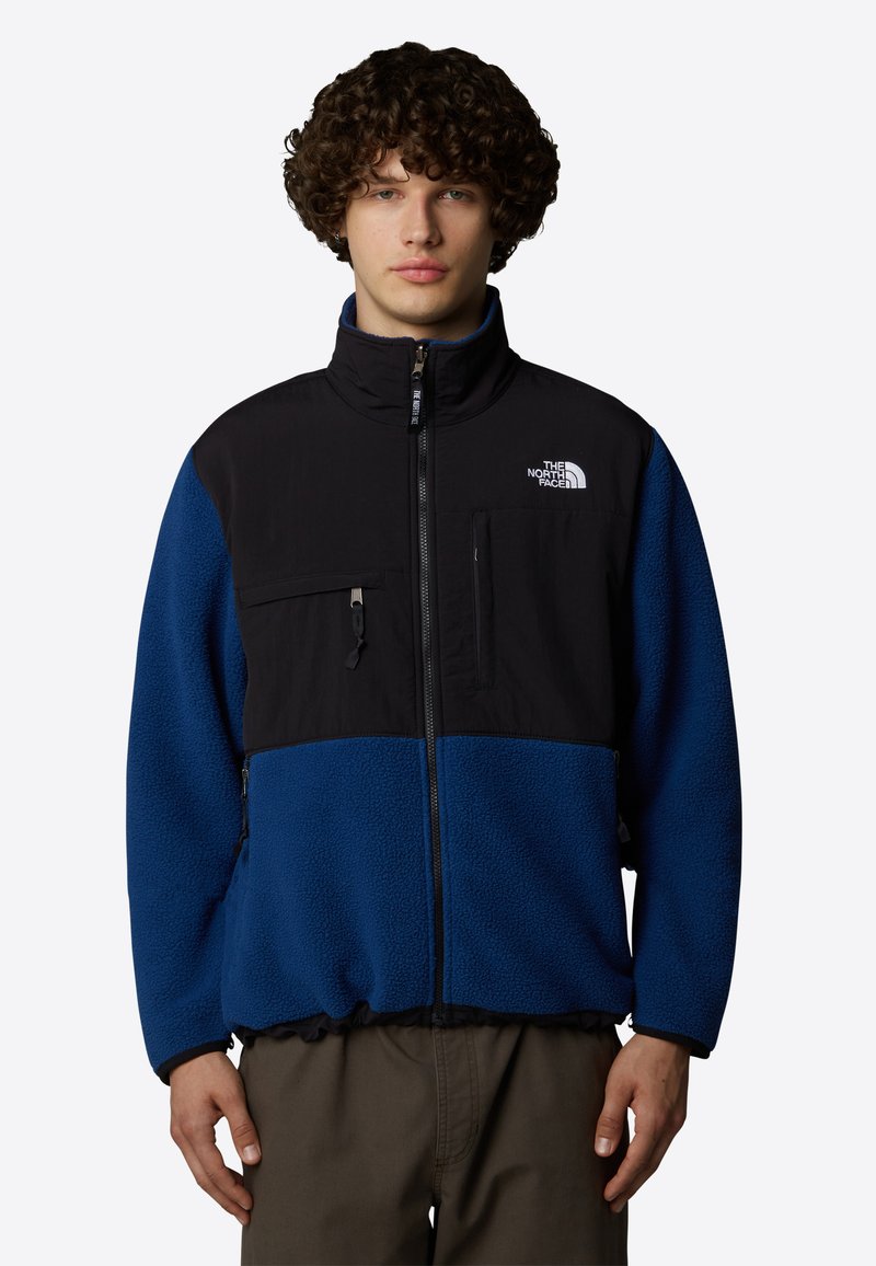 Czarna i niebieska polarowa kurtka z wysokim kołnierzem; kieszeń na zamek błyskawiczny na piersi; logo po lewej stronie klatki piersiowej; teksturowany materiał; elastyczny pas i mankiety.