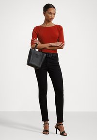 Rode fitted top, zwarte skinny jeans, zwarte riem met textuurelementen, zwarte handtas met logo en zwarte sandaaltjes met straps. Eenvoudige achtergrond.