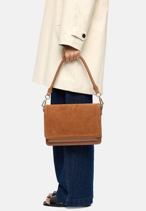 Osoba w beżowym płaszczu i ciemnych dżinsach trzyma brązową zamszową torebkę z rączką na górze na jednolitym tle.