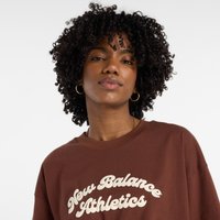 T-shirt oversized castanho com "New Balance Athletics" em fonte creme. Cabelo cacheado, brincos de argola e design minimalista. Textura de tecido suave.