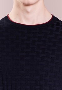 Gros plan sur le cou et les épaules d'une personne portant un pull sombre à texture avec un col rond et une fine bordure rouge.