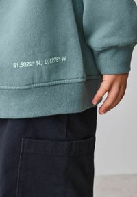 Világoskék-zöld pulóver texturált anyaggal, amelyen fehér koordináták szerepelnek: "51.5072° N, 0.1276° W" a szegélynél. Sötét nadrággal viselve.