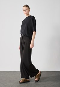 Camiseta negra de corte holgado con mangas cortas; pantalones negros de pierna ancha; sandalias deslizantes con estampado animal y suela plana.
