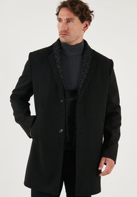 Abrigo de lana negro con solapas de muesca, dos botones y bolsillos frontales. El cuello interior presenta un patrón geométrico en gris.